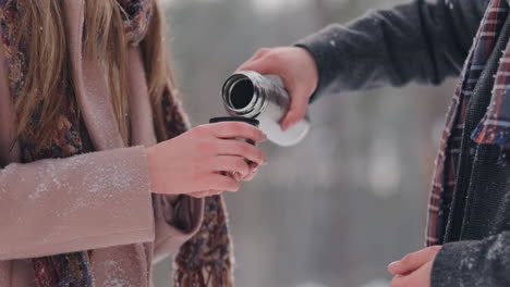 A-loving-couple-man-and-woman-in-the-winter-forest-drinking-tea-from-a-thermos.-Stylish-man-and-woman-in-a-coat-in-the-Park-in-winter-for-a-walk.-Slow-motion